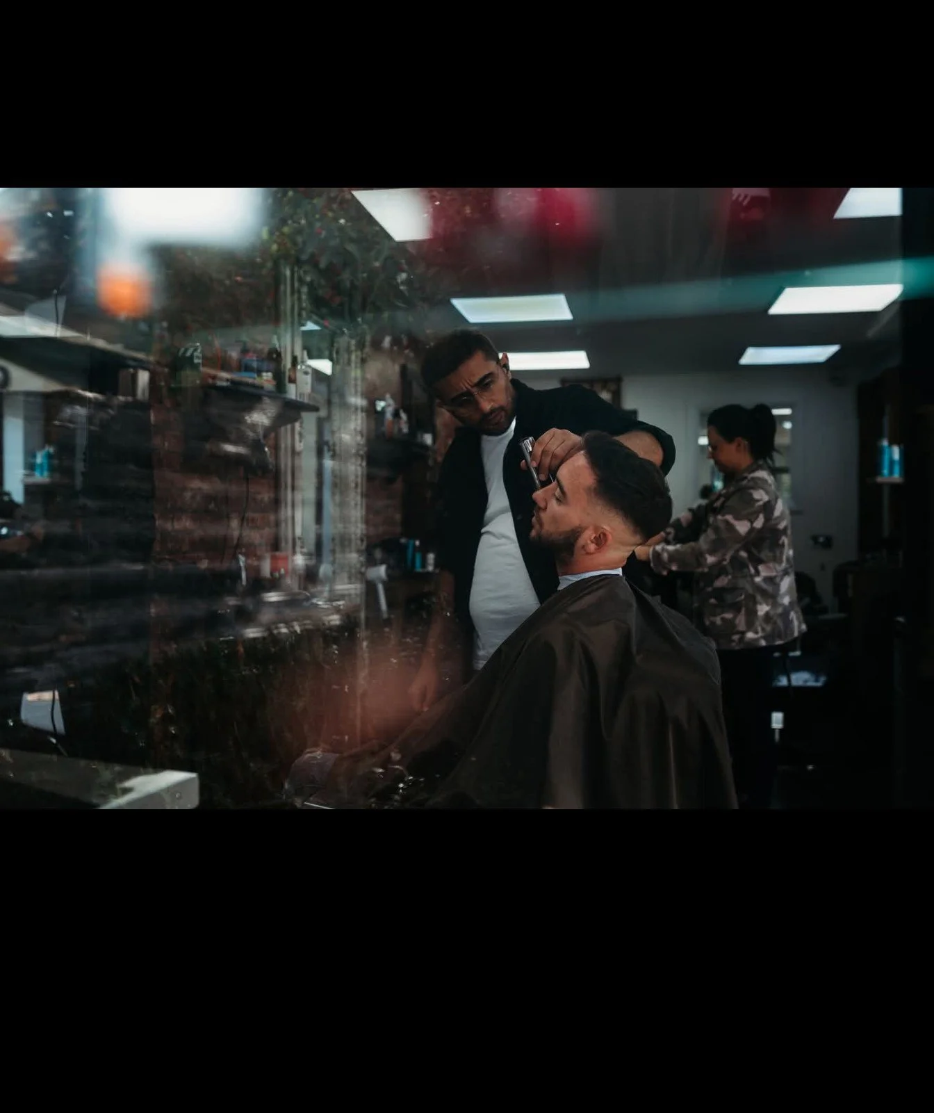 A man getting a haircut at a barbershop, viewed through a glass window.