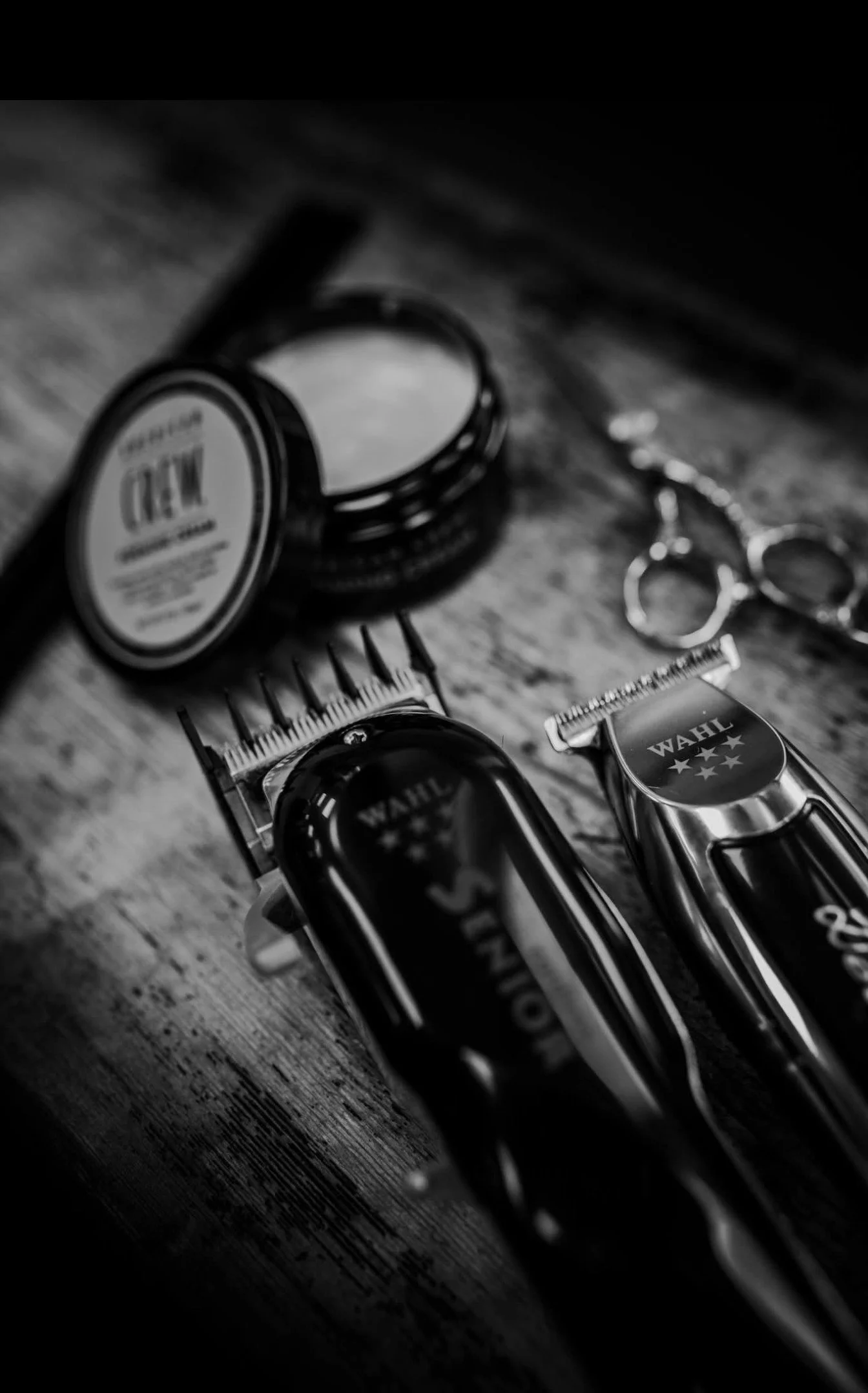 Black and white photograph of hair grooming tools, including electric clippers and scissors, placed on a dark wooden surface.
