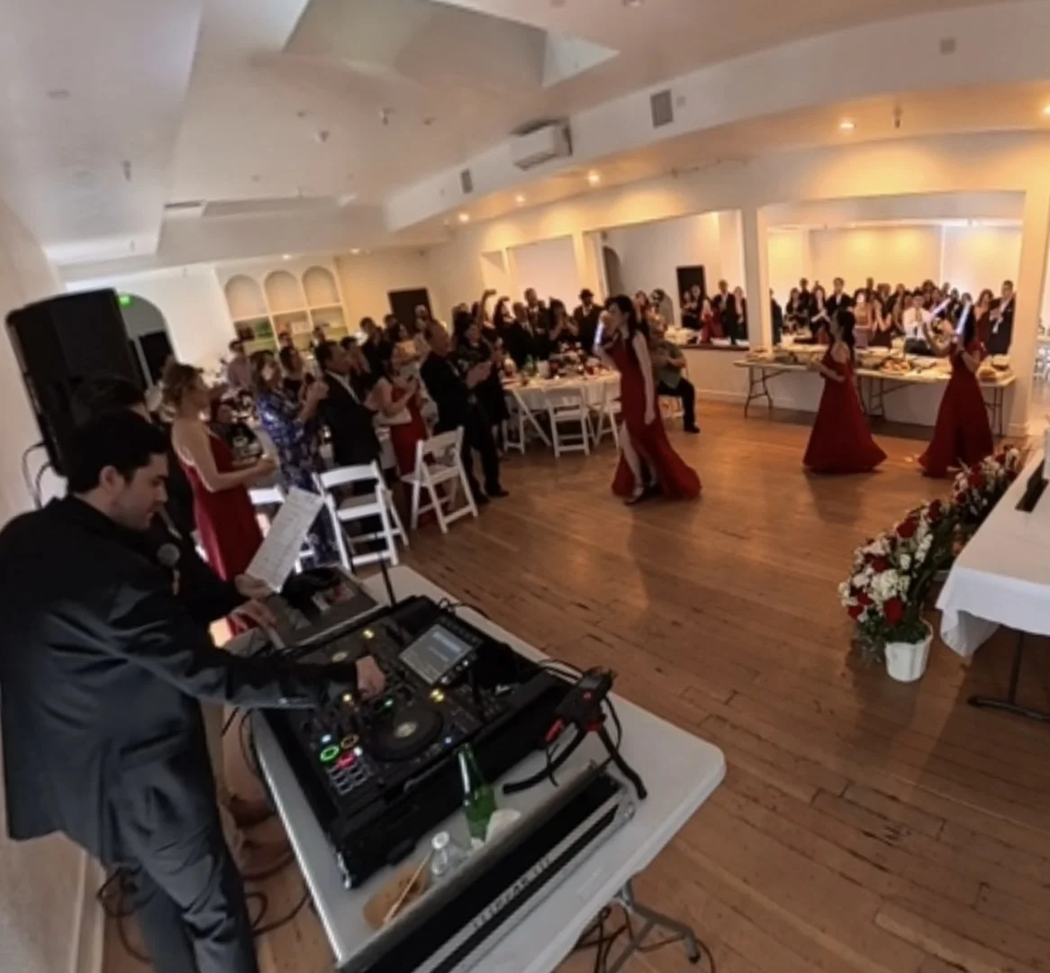 Wedding Dj Cliff Mertens on the mic at a wedding reception as the bridal party makes their entrance in red dresses before a standing crowd.