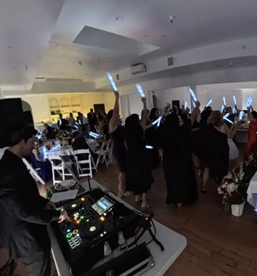 Wedding DJ Cliff Mertens performing for a packed dance floor with guests waving glow sticks near decorated tables and floral arrangements.