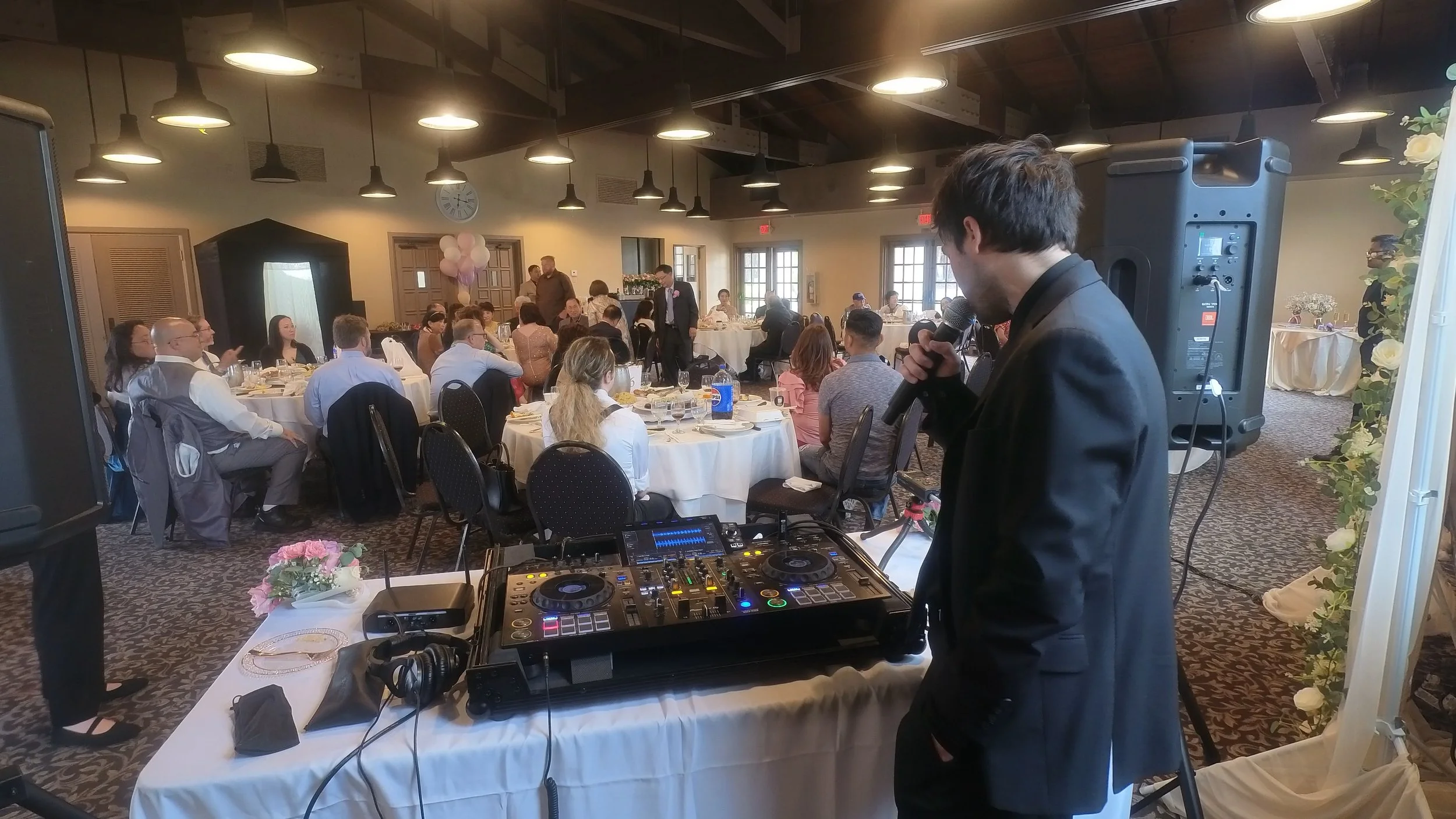 Wedding DJ and MC Cliff Mertens on the microphone at his set up as guests dine at a wedding reception.