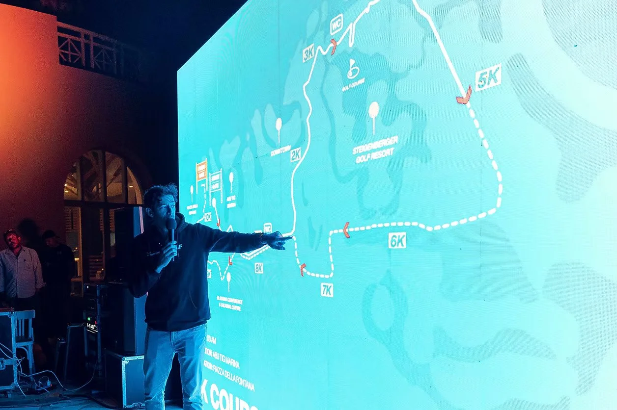 A man holding a microphone and pointing at a large digital map of a running course displayed on a screen. The map shows different running distances marked as 2K, 3K, 5K, 6K, 7K, and 8K, with a route through a park or golf course area.