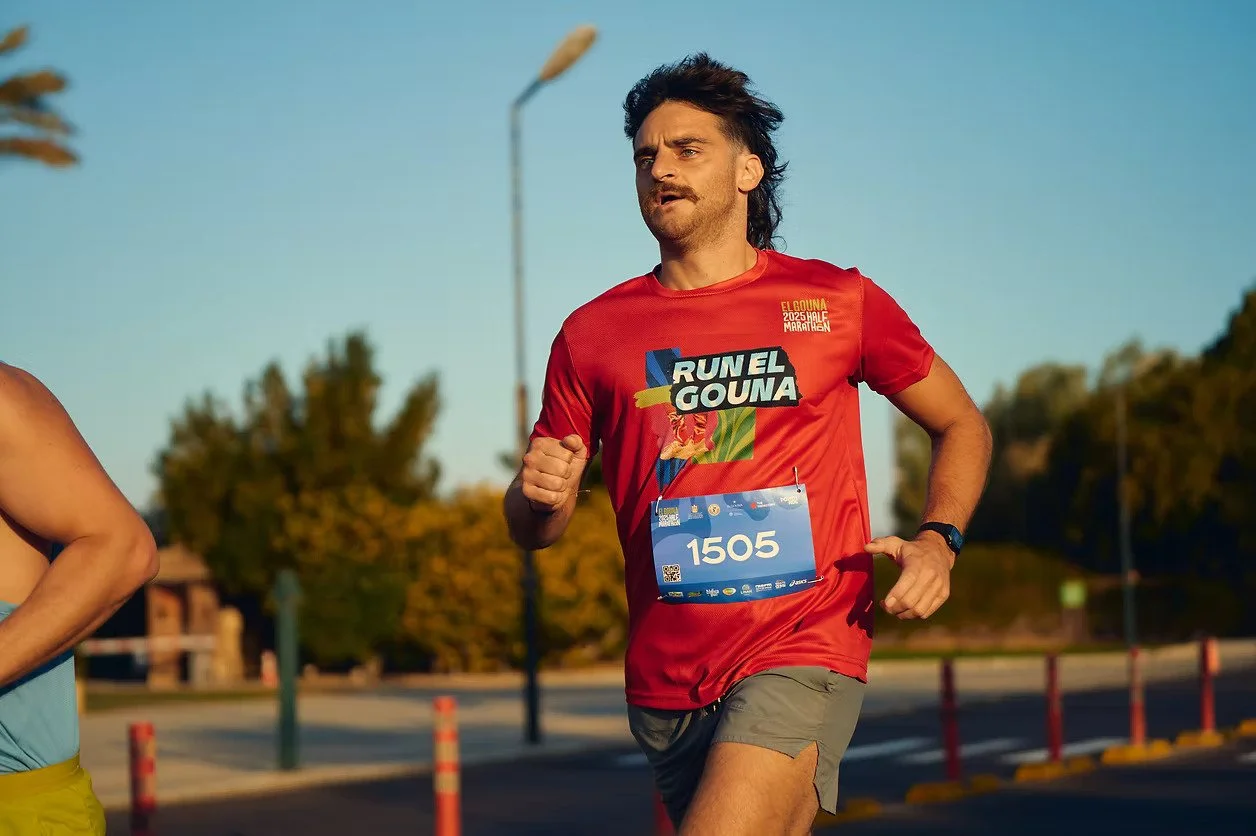 A man running in a marathon wearing a red shirt with the text 'RUN EL GOUNA' and a race bib number 1505, on a road in warm weather with trees and streetlights in the background.