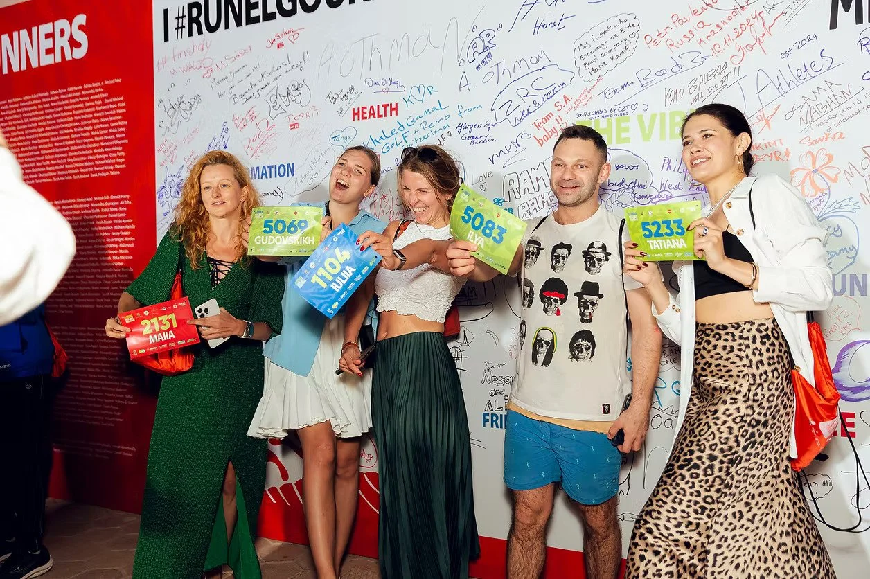 Group of five diverse young adults standing in front of a white wall with colorful signatures, holding colorful card signs with numbers and names, smiling and posing for the photo at an event.