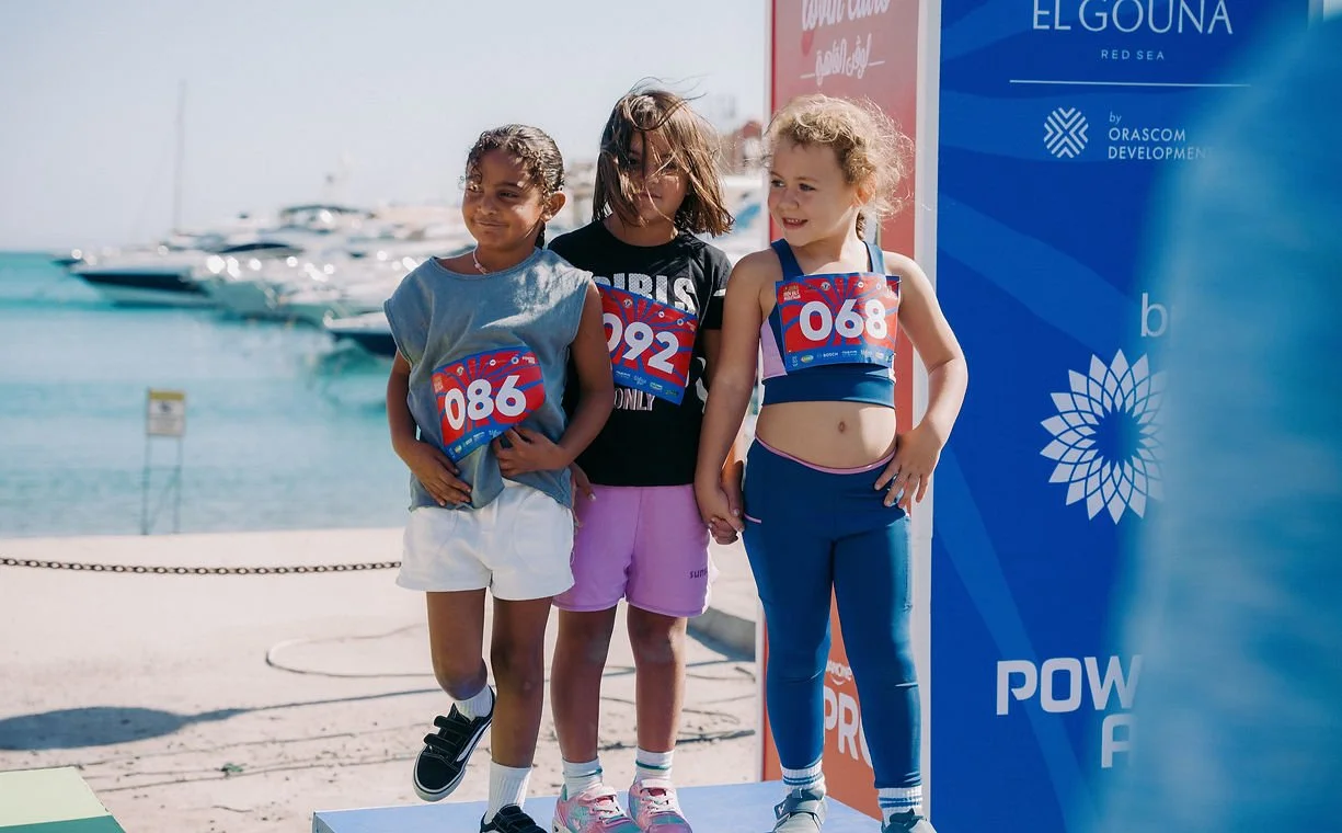 Three young girls standing together on a beach near yachts, each wearing medals and numbered bibs, during a race event.
