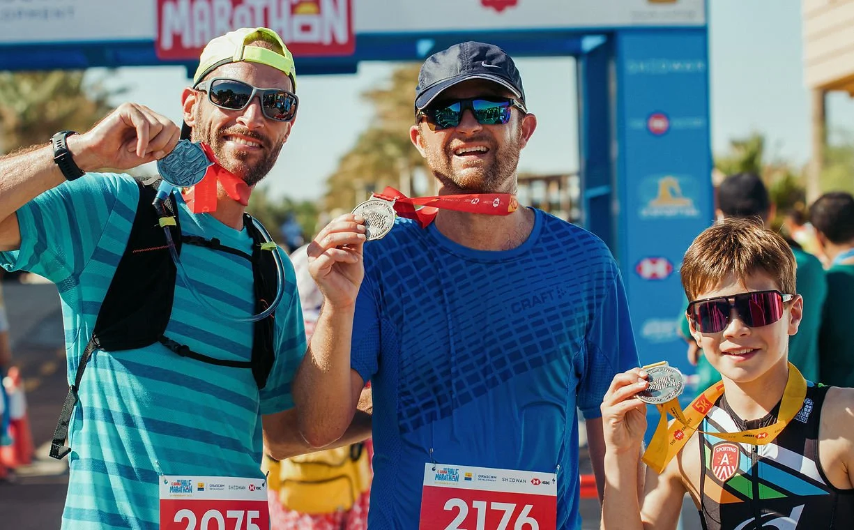 Three runners displaying their marathon medals after finishing the race, a man with sunglasses and a cap, a man in a blue shirt with a cap, and a young boy with sunglasses all smiling.