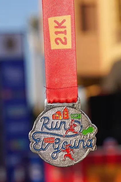 A silver finish tourism medal for the Rim 2 Eryri race, featuring icons of a mountain, a cyclist, and a swimmer, attached to a red ribbon with '21K' printed on it.