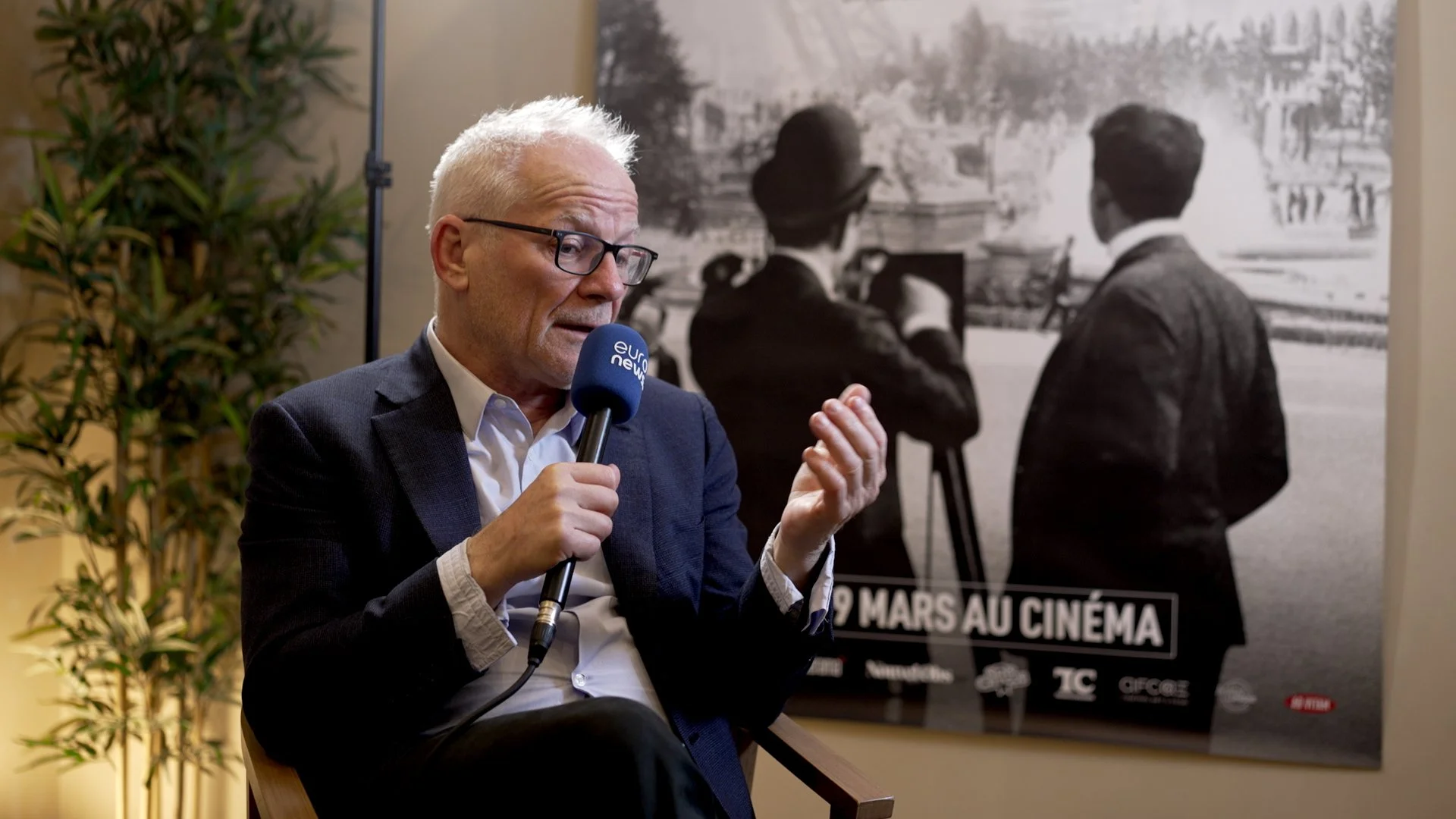 An interview with Thierry Frémaux. Behind him is a large black-and-white poster showing two people with their backs turned, looking at a cityscape, with some text at the bottom.
