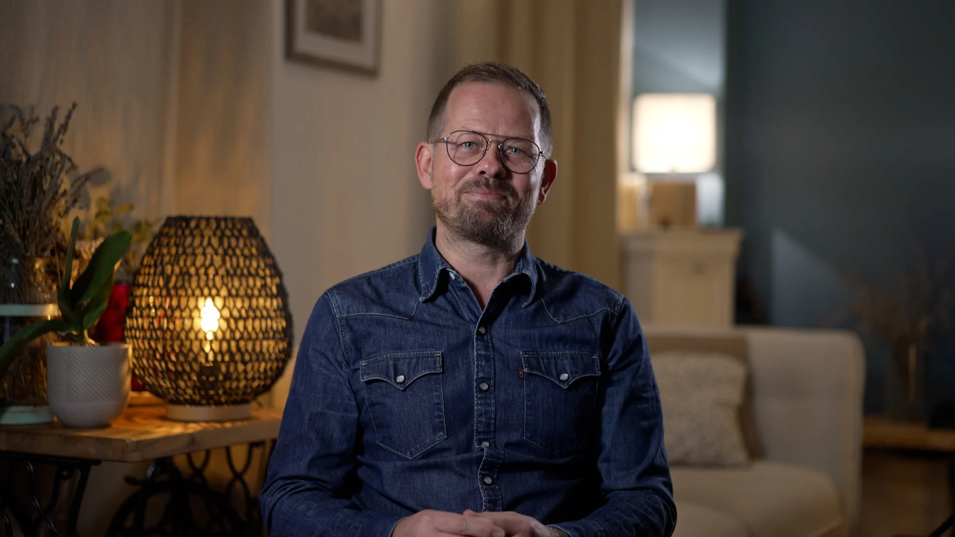 Photo of videographer Joseph Allen wearing a denim shirt, sitting in a cozy living room with warm lighting and decor.
