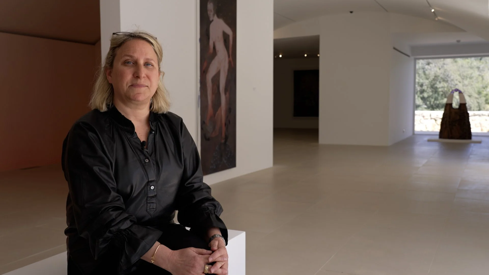 Alon Pardo, wearing a black button-up shirt, sitting in an art gallery or museum. Behind her, white walls feature artwork, and large windows reveal trees outside.
