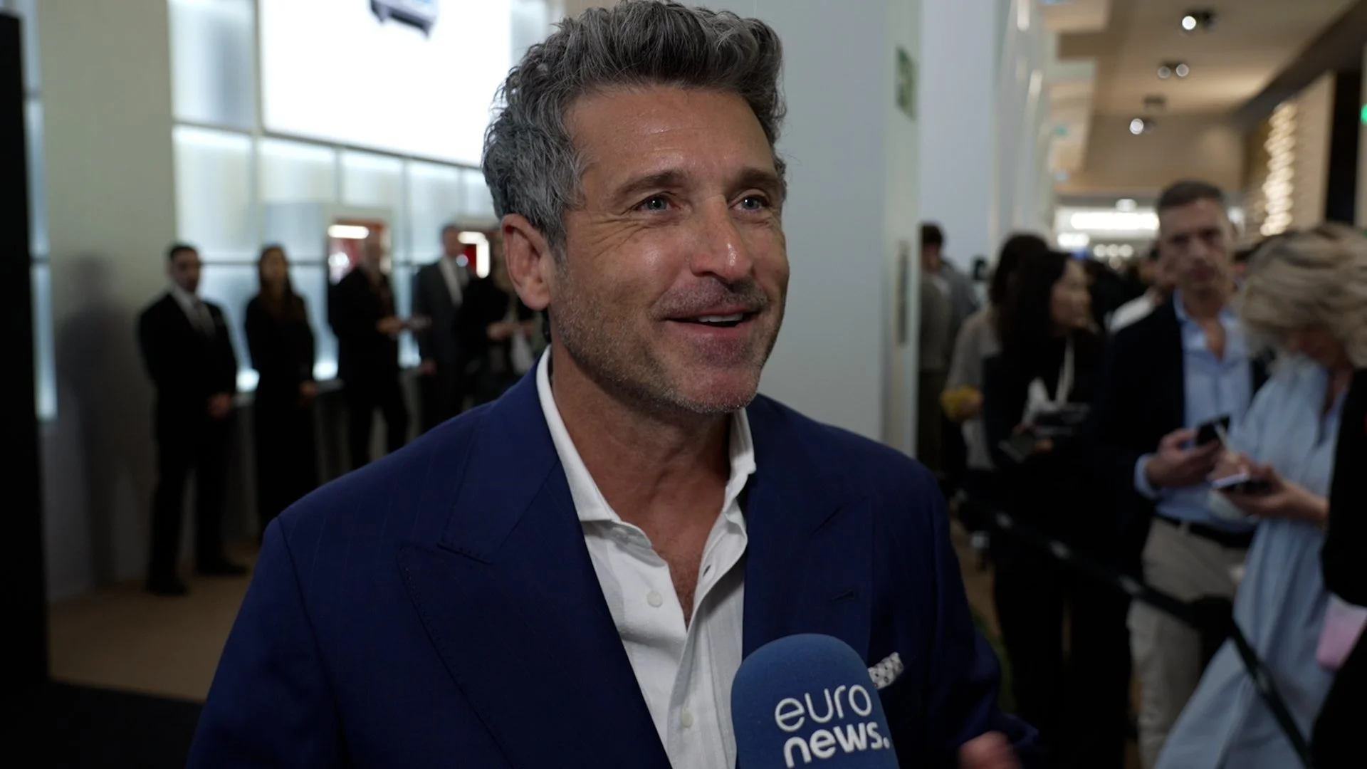 Patrick Dempsey in a blue suit being interviewed by a EuroNews reporter at a Watches and Wonders in Geneva, with people in business attire in the background.