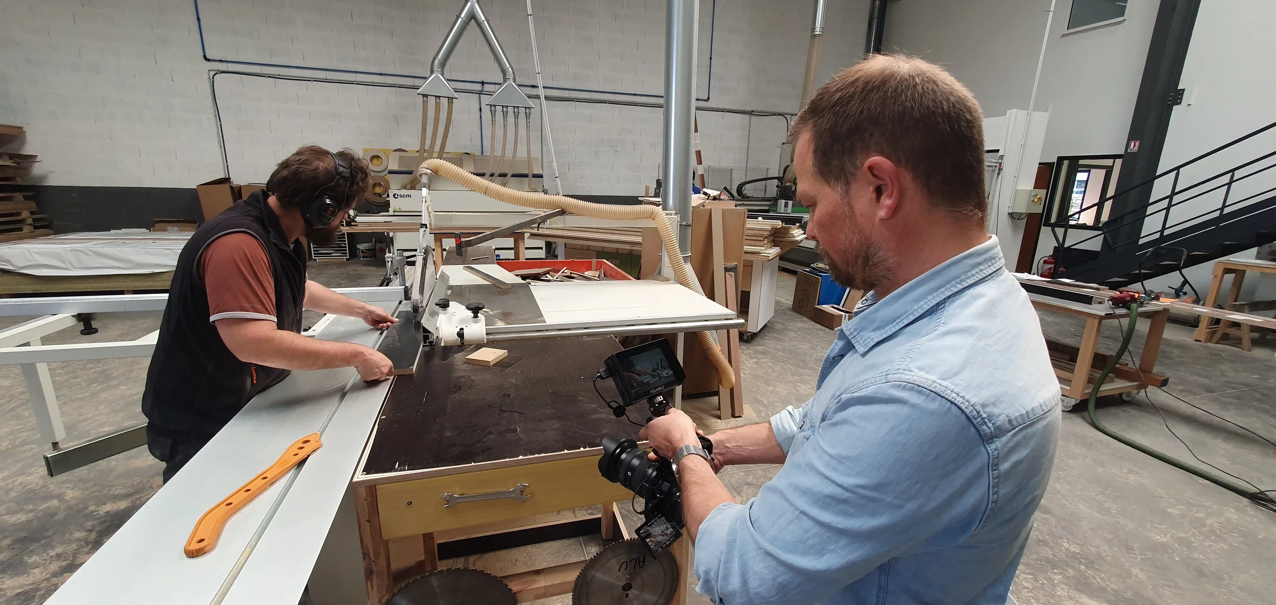 Joseph Allen filming in a woodworking shop, one operating a machine and the other filming or photographing the process, surrounded by woodworking tools and materials.