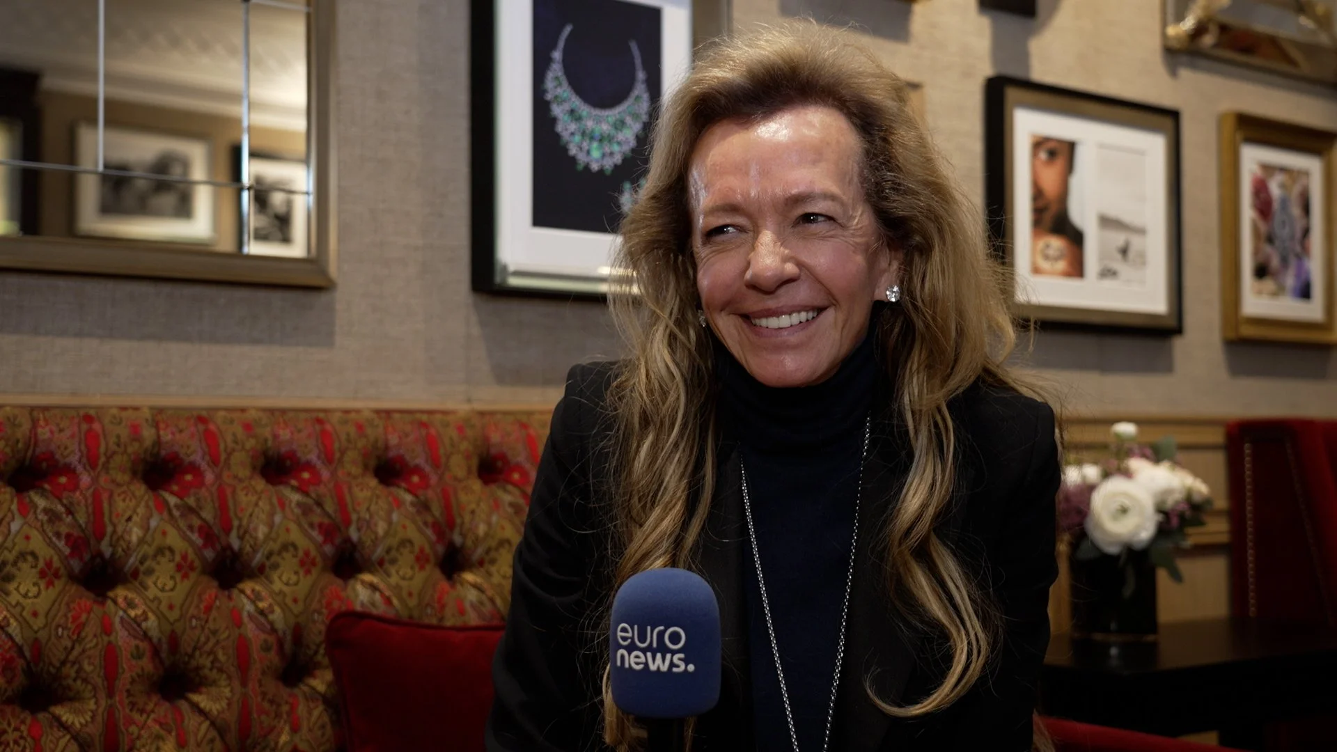 Caroline Scheufele smiling, sitting on a patterned red and gold upholstered sofa in a room with framed artwork on the wall behind her. She is wearing a black turtleneck and a jacket, and is speaking into a blue EuroNews microphone.