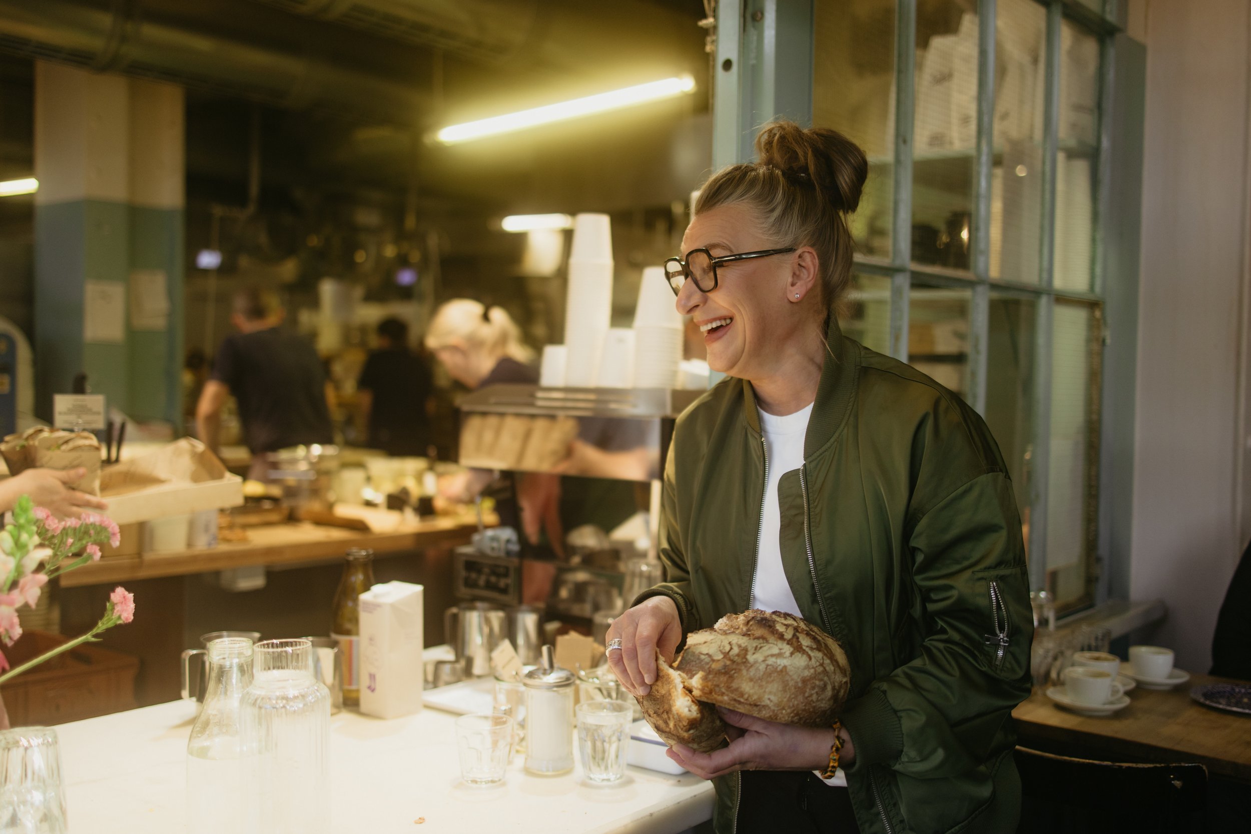 Frau mit Brille und Knotenkopf arbeitet in einem Café, hält ein großes Stück Brot in den Händen, im Hintergrund erscheinen weitere Mitarbeiter, an der Theke sind Gläser und Teller sichtbar.