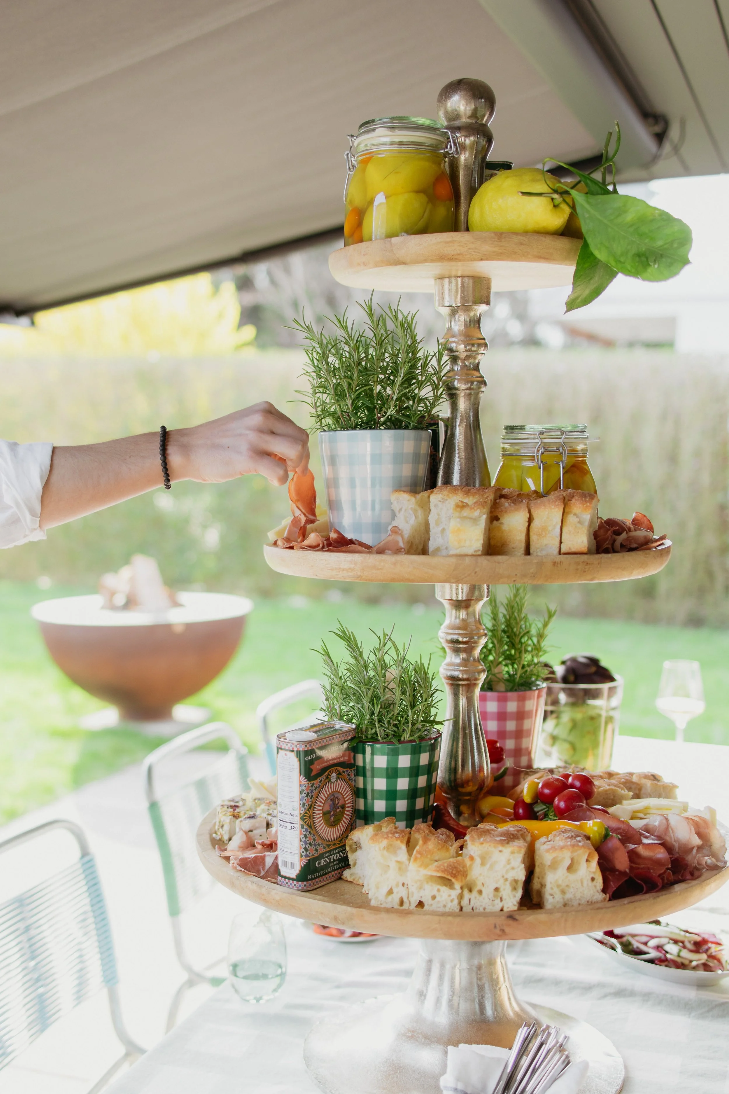 Ein mehrstöckiges Buffet mit verschiedenen Speisen, Getränken, frischen Früchten, Broten, Oliven und einer Topfpflanze auf einem gedeckten Tisch im Freien, bei hellem Tageslicht.