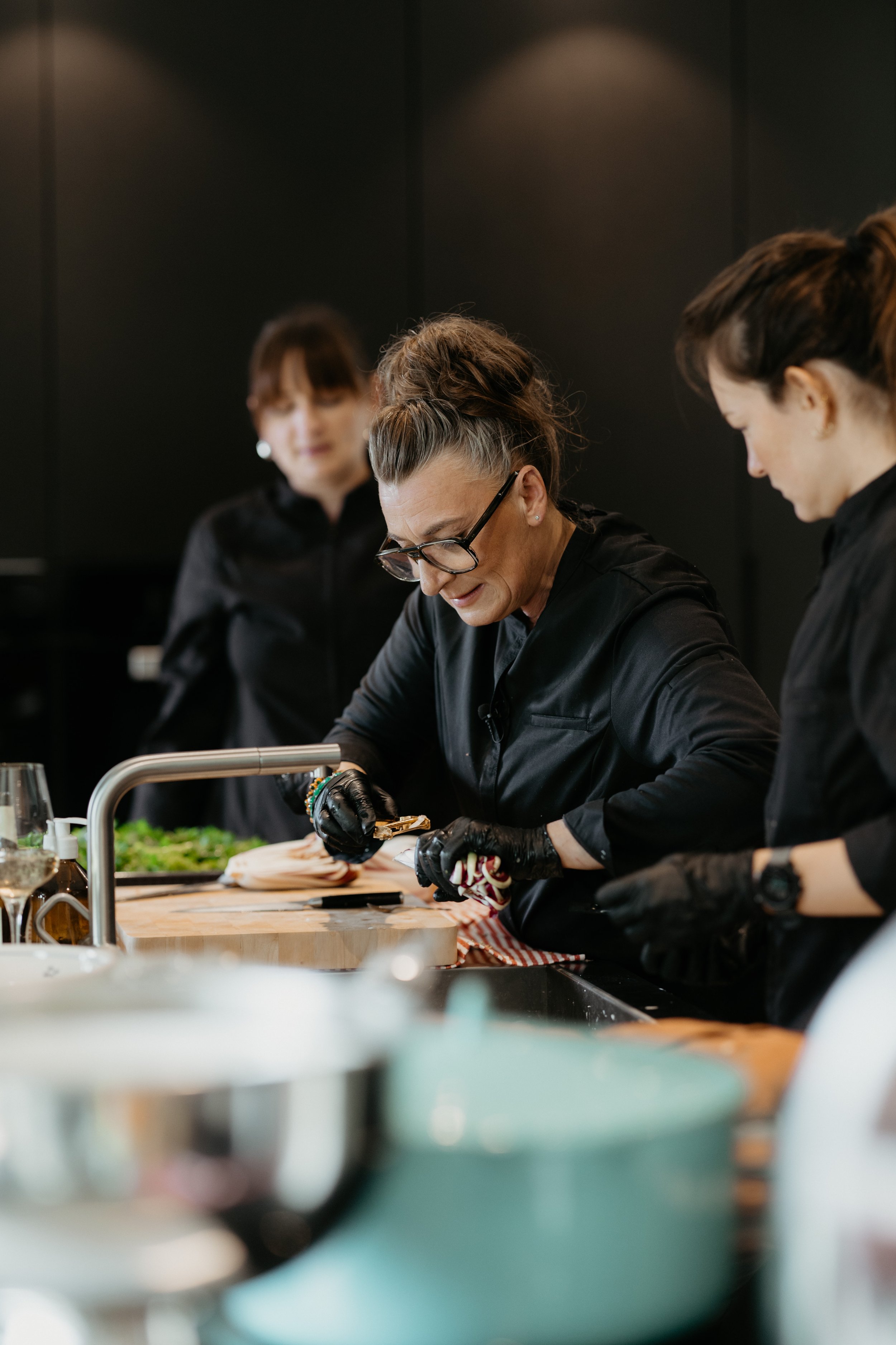 Drei Frauen in schwarzen Kochuniformen bereiten Gerichte in einer modernen Küche zu. Im Vordergrund sind blinde Flecken, während die mittlere Frau mit Brille sorgfältig an einem Gericht arbeitet. Die beiden Frauen im Hintergrund beobachten oder warten.