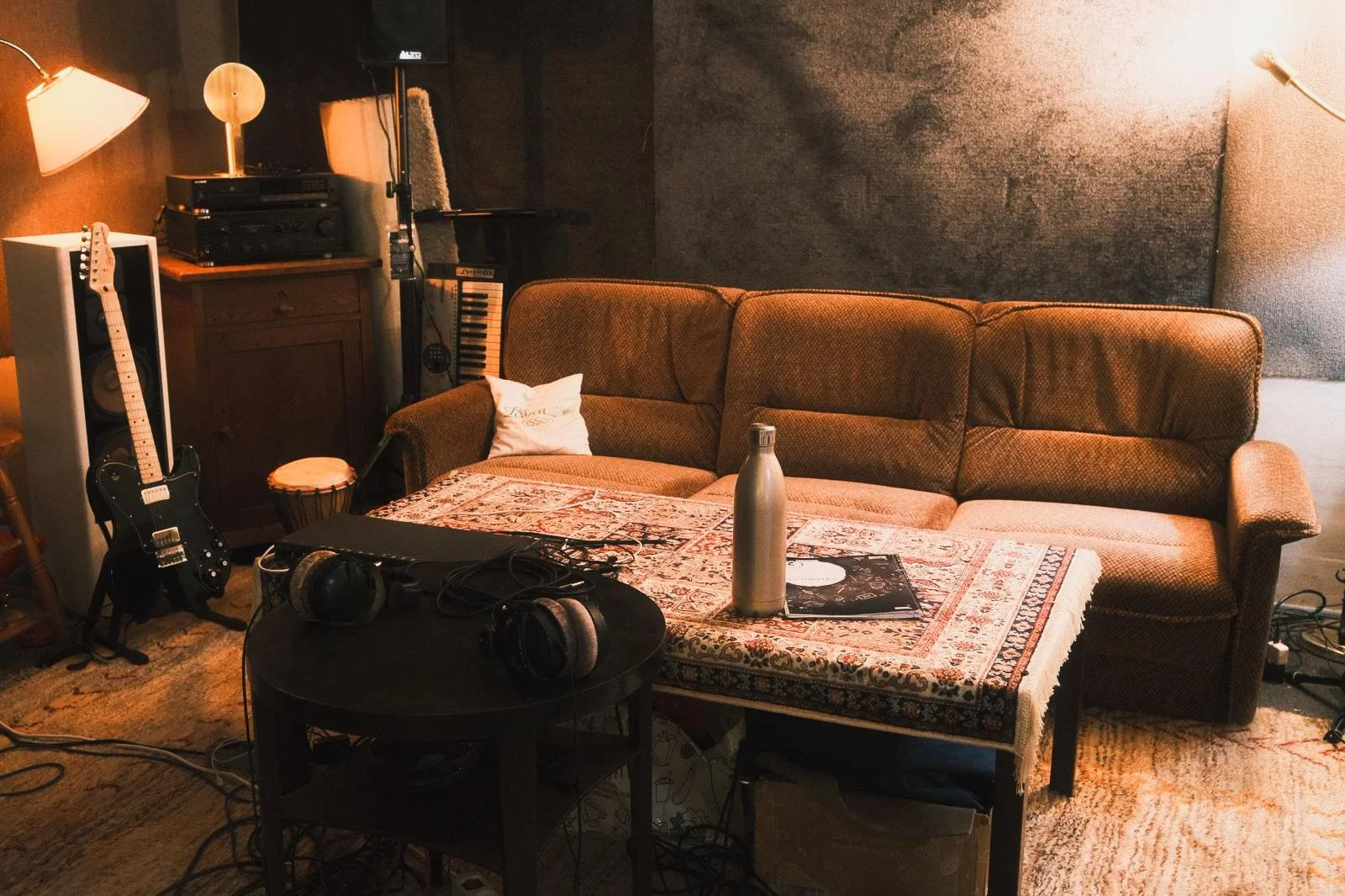Musikzimmer mit braunem Sofa, einem Tisch mit einem Wasserflasche, Kopfhörern, einem Collge-Block, sowie Gitarre, Verstärker, elektronisches Keyboard und Lautsprechern.
