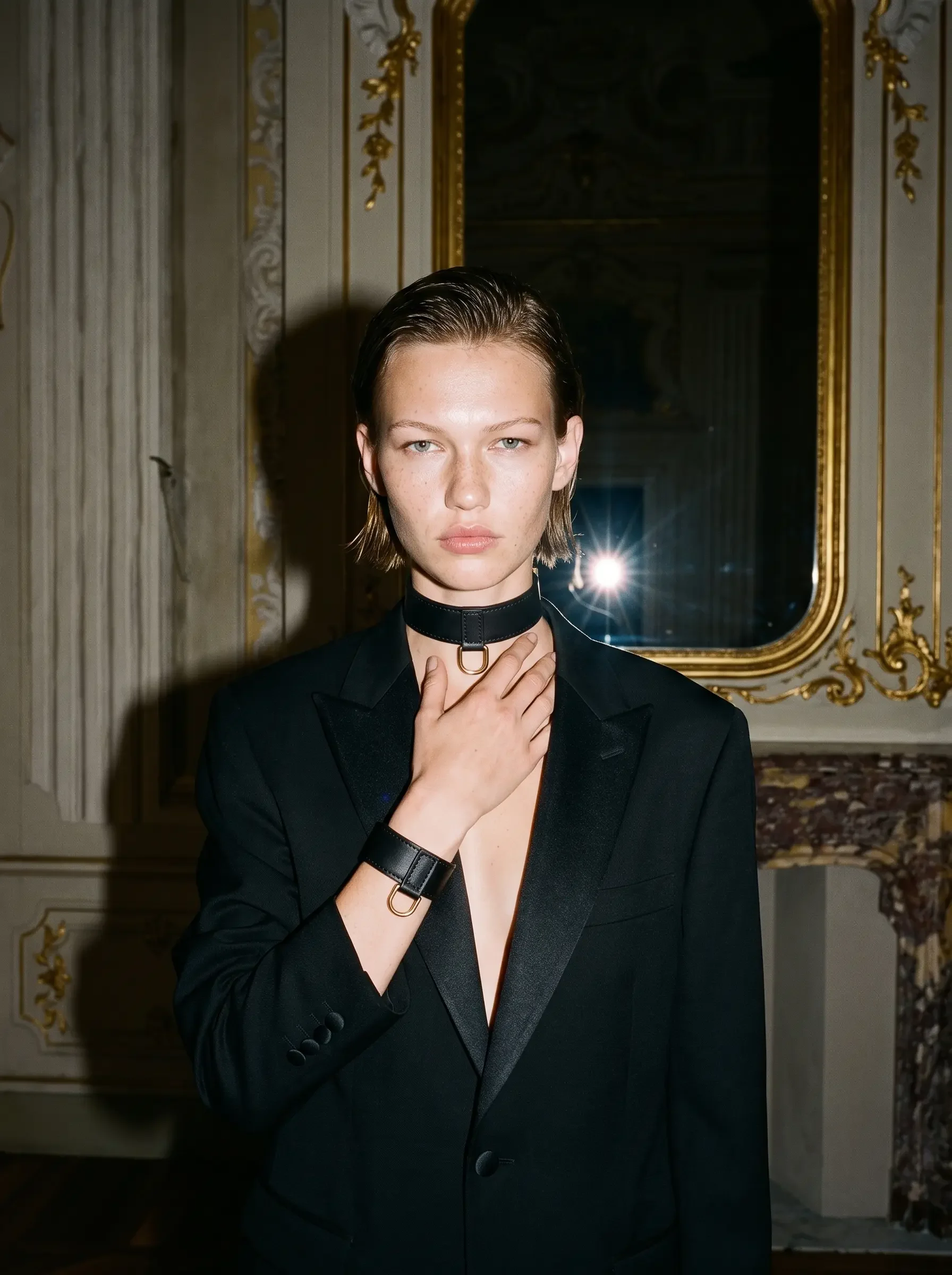 Sainte Sve choker and wrist bracelet in black nappa lambskin with 24K gold-plated brass hardware, worn by a model in a black blazer before a gilded mirror, editorial image