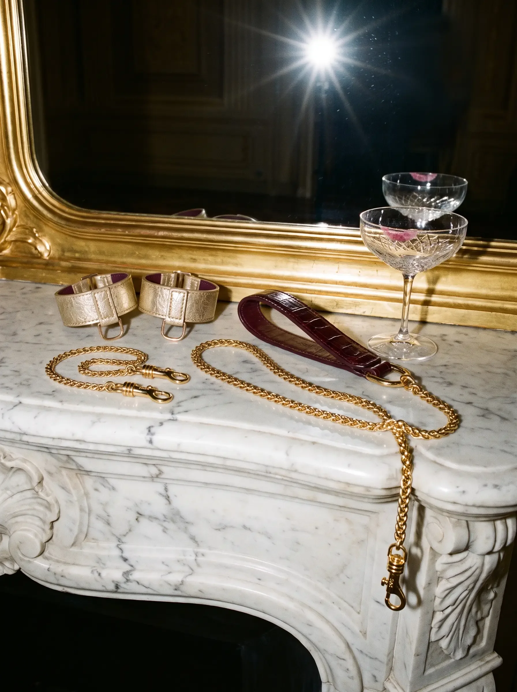 Sainte Sve wrist bracelets in gold laminated lambskin, Handcuff Chain and burgundy Leash Croc on a marble mantelpiece beneath a gilded mirror, editorial image