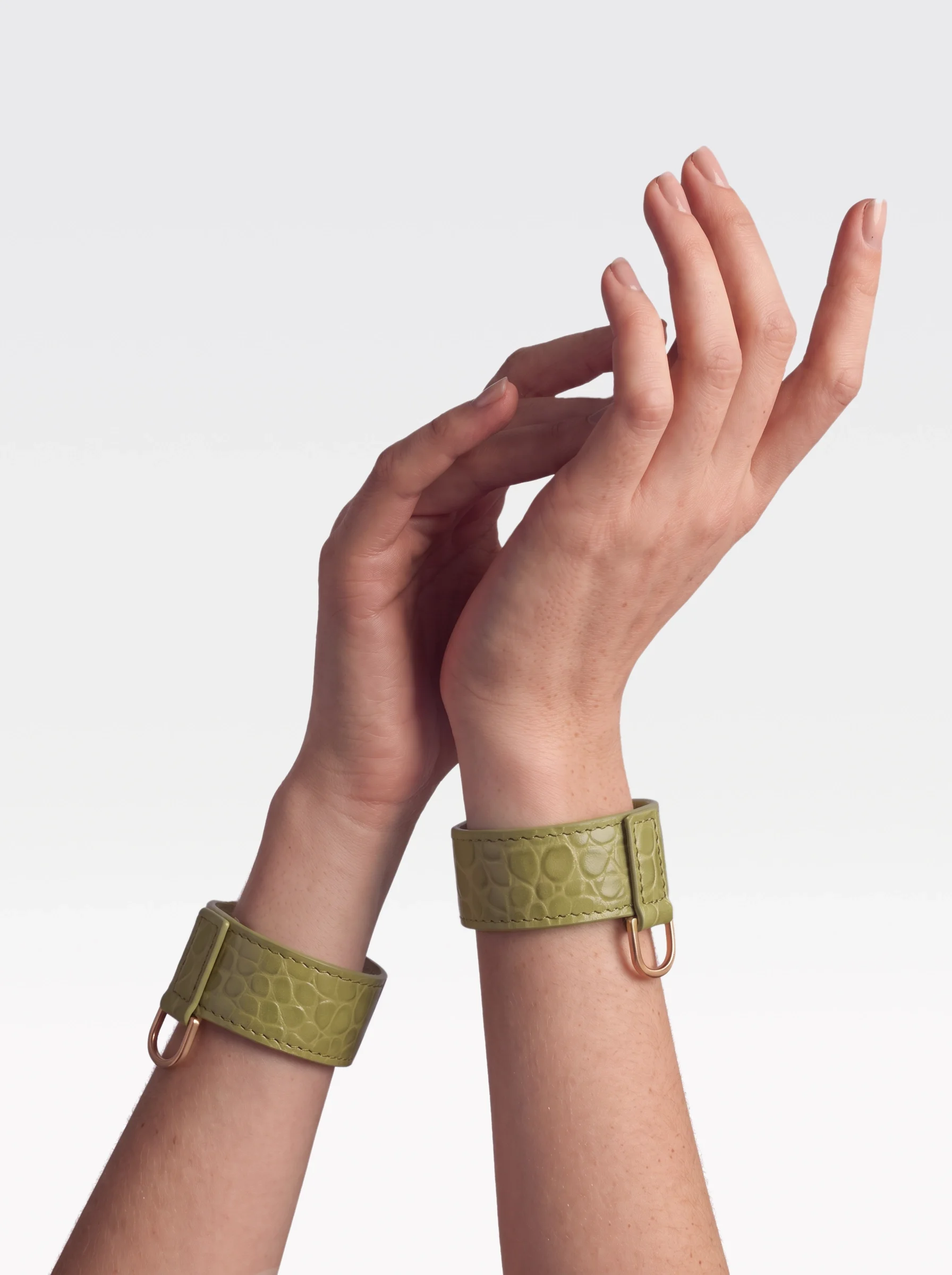 Sainte Sve Wrist Bracelets Croc in pistachio crocodile-embossed lambskin with 24K gold-plated brass D rings, worn on raised wrists, editorial image