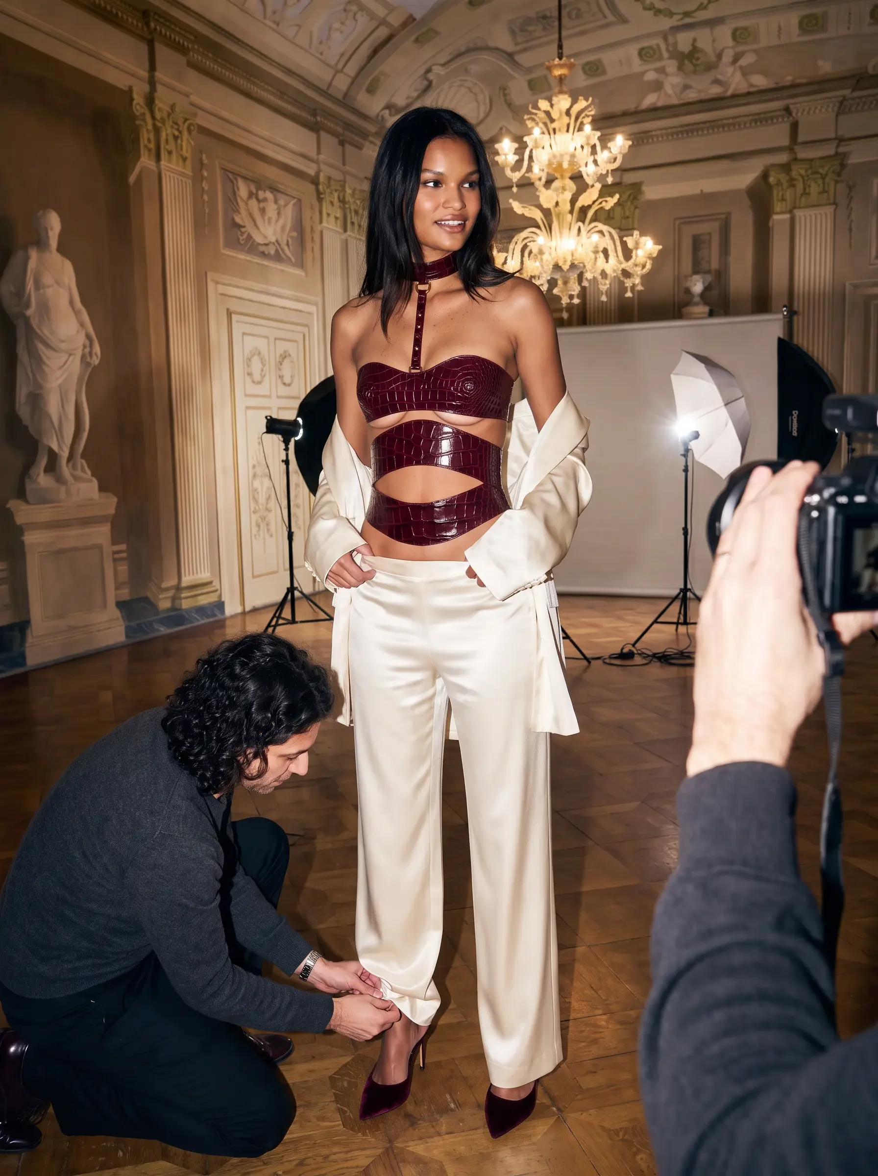 Sainte Sve Corset V.N.V in burgundy crocodile-embossed lambskin with 24K gold-plated brass hardware, worn with cream trousers and a white jacket on a photo shoot set, editorial image