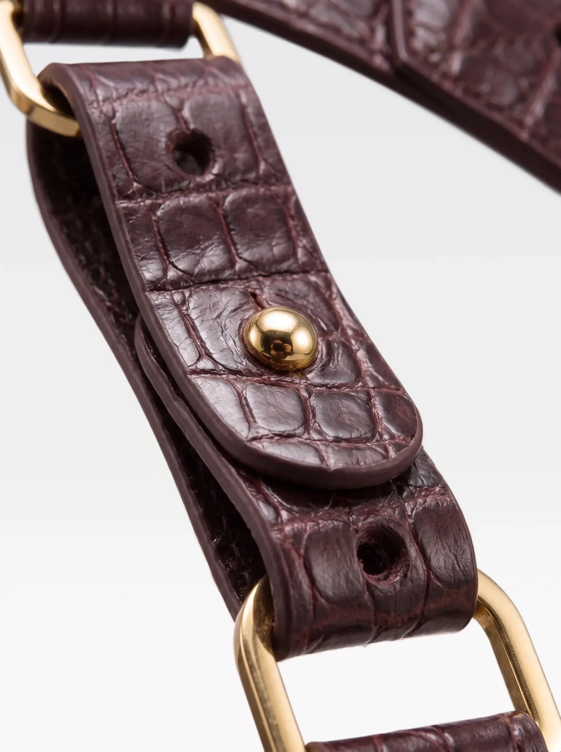 Close-up of the 24K gold-plated brass connector rivet and D ring on the burgundy crocodile-embossed lambskin strap of the Sainte Sve Belt Plus Croc