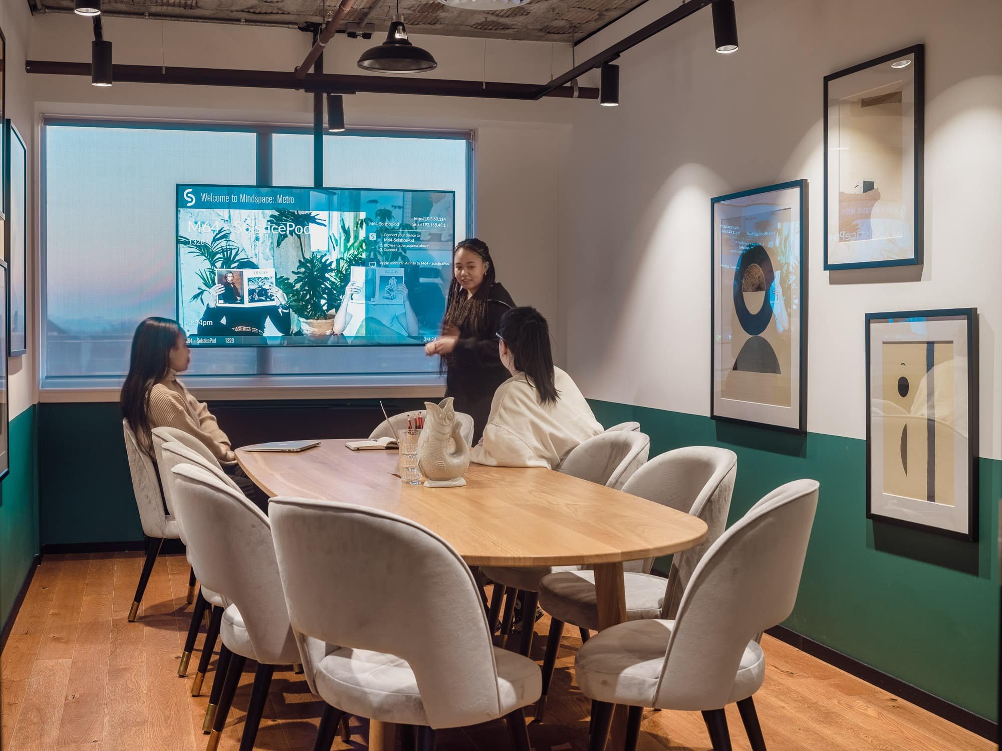 A group of four women sitting around a wooden table in a modern art gallery or museum, with colorful abstract paintings on the walls and a large digital screen displaying information in the background.