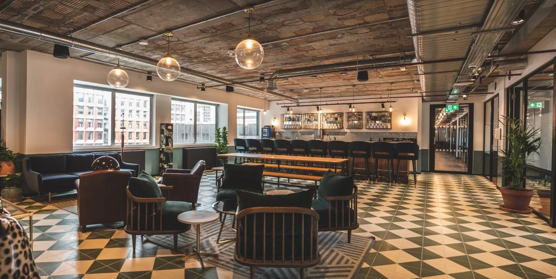 Modern lounge with black and green seating, wooden tables, decorated ceiling, and checkered floor, featuring large windows, indoor plants, and a bar area in the background.