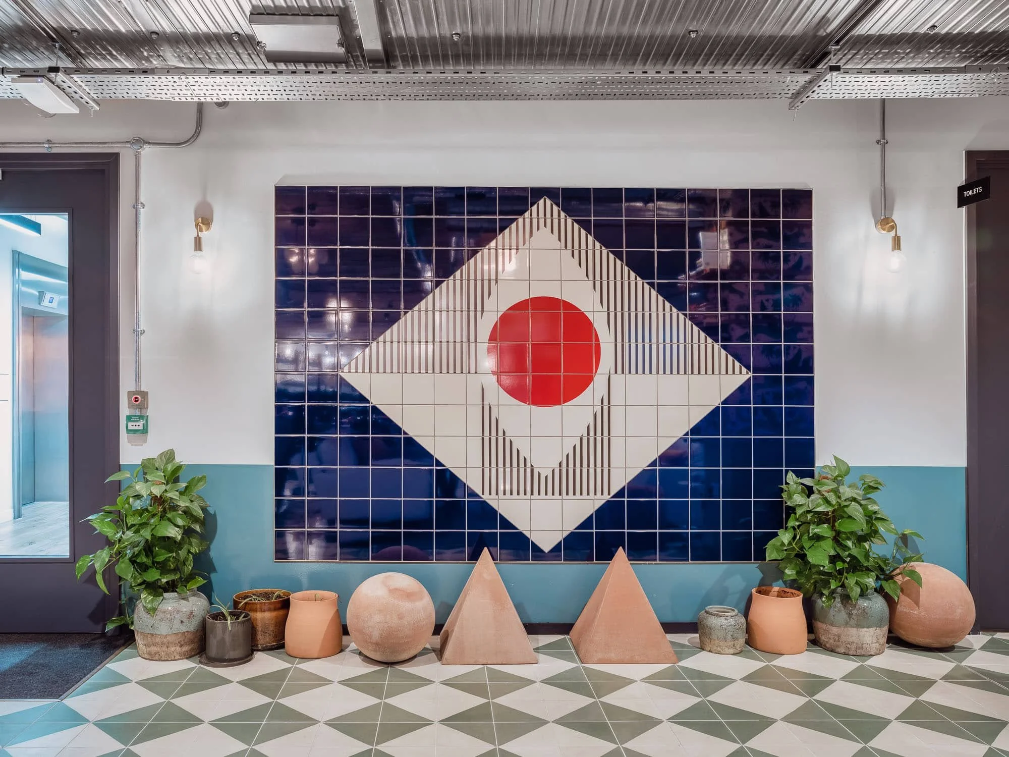 Decorative wall with dark blue tiles, a geometric pattern with a red circle in the center, six potted plants at the base, and a tiled floor with a diamond pattern.