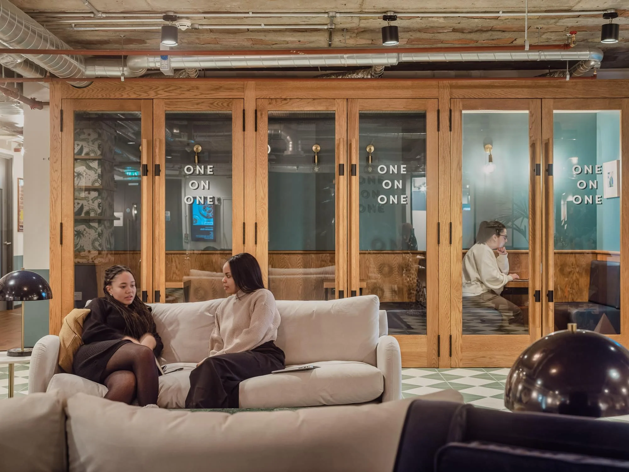 People sitting on a beige sofa in a modern office lounge, with a wooden partition and glass doors behind them. Two women are engaged in conversation, while a woman with glasses and a white sweater is seated at a desk to the right.