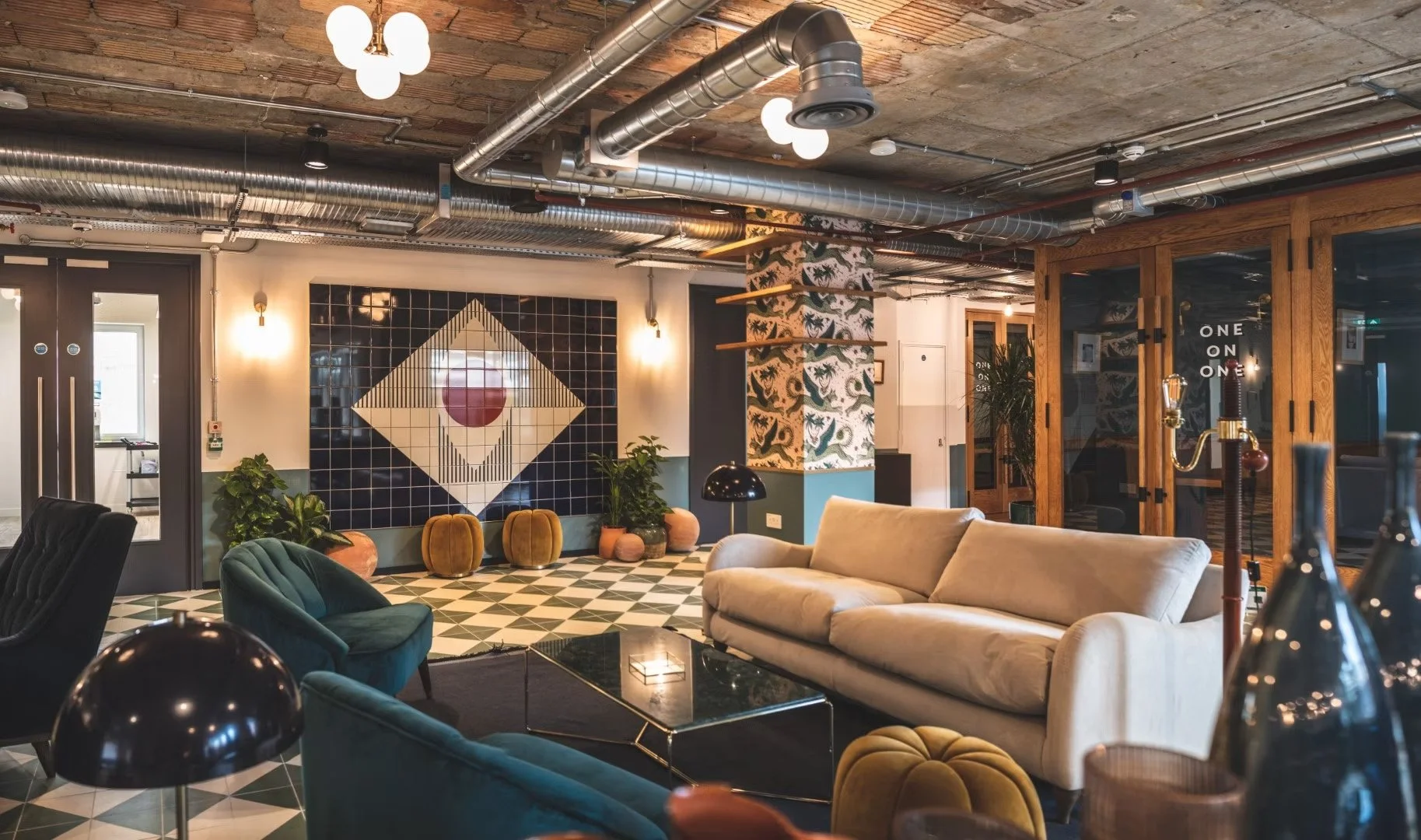 Interior of a modern lounge with plush sofas, plants, and decorative wall art, featuring exposed ceiling ducts and patterned tile flooring.