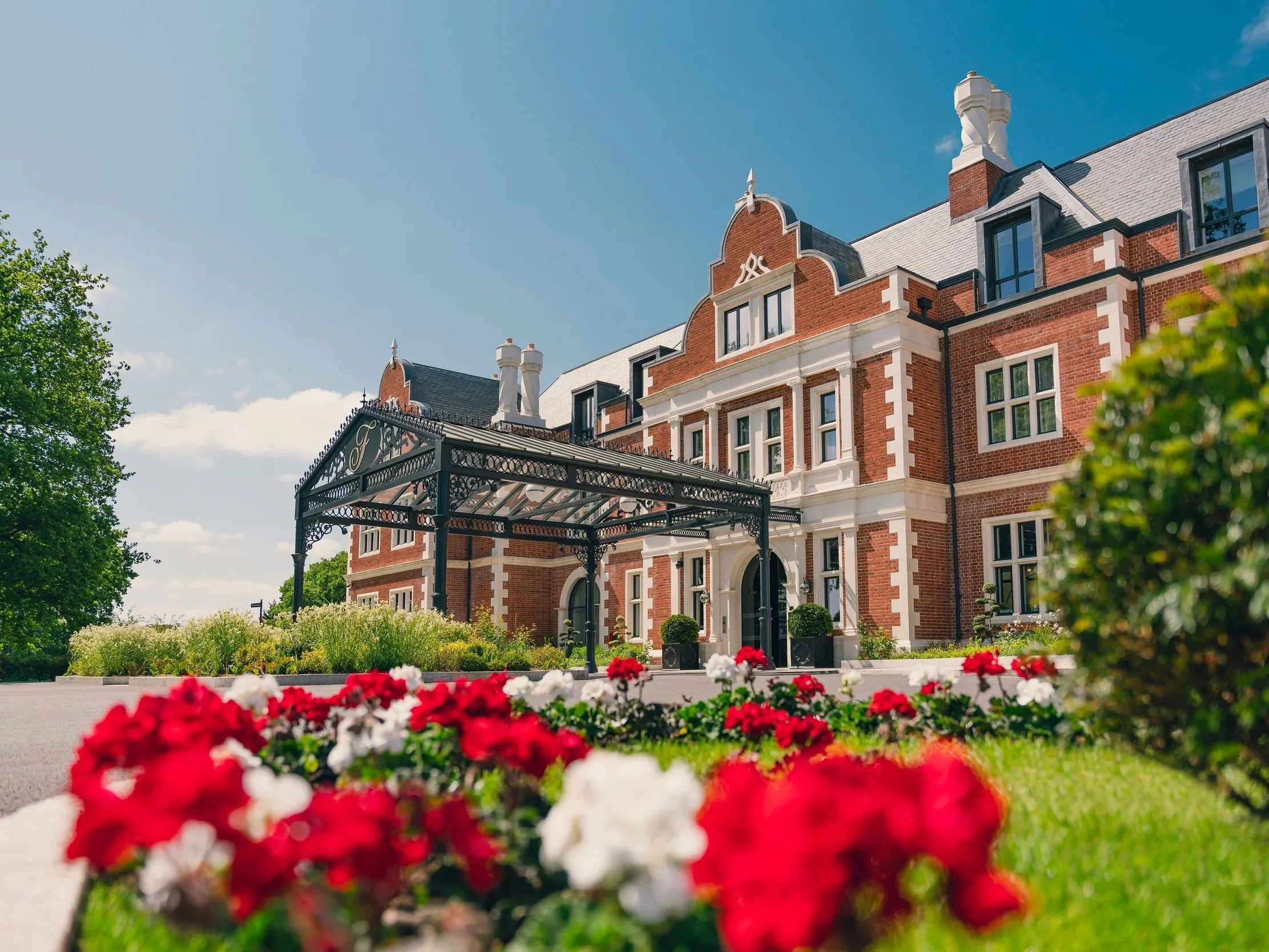 A large red brick building with white accents and a decorative wrought-iron awning over the entrance, surrounded by green trees and a garden bed with red and white flowers, under a blue sky.
