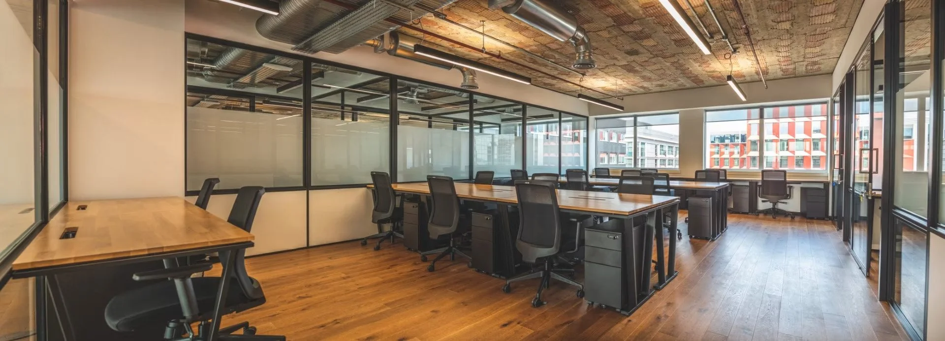 Modern empty office space with large windows, glass walls, wooden floors, and multiple desks with black office chairs.