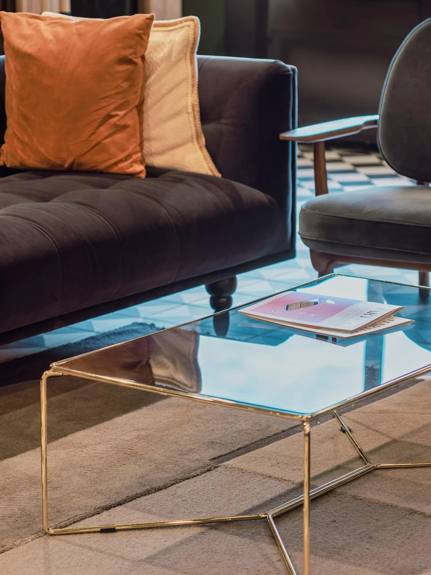Living room with a black velvet sofa, orange and beige pillows, a glass coffee table with a gold frame, a magazine on the table, a gray armchair, a rug with a striped pattern, and hardwood flooring.