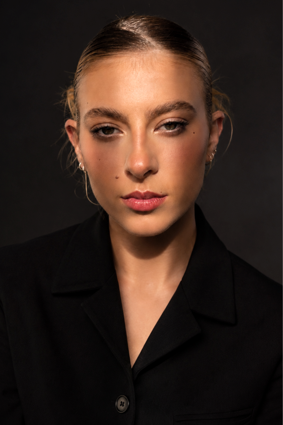 Portrait d'une jeune femme avec des cheveux blonds tirés en arrière, portant un uniforme noir, contre un fond sombre.