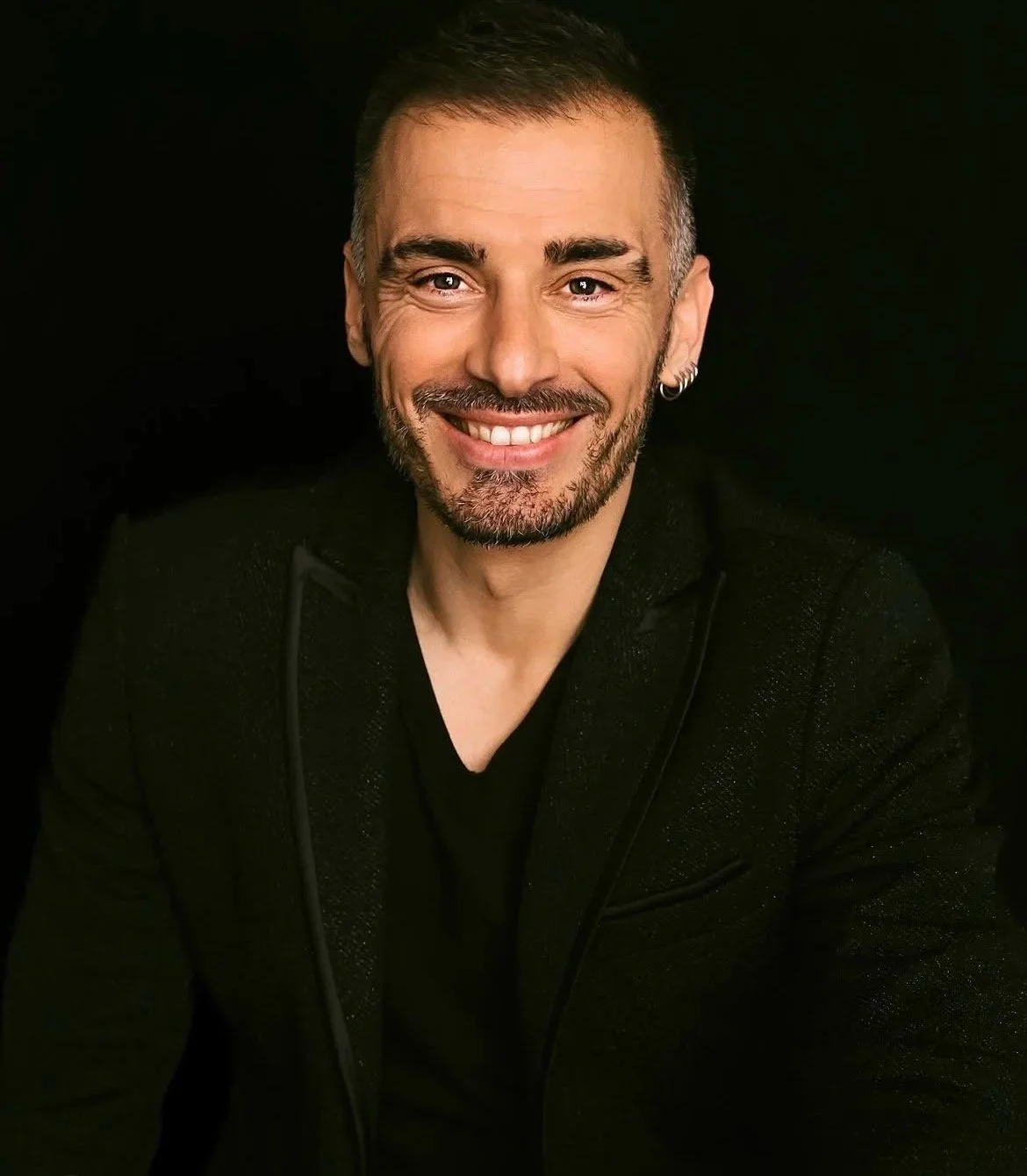 Un homme souriant avec une barbe, portant un blazer noir, sur fond noir.