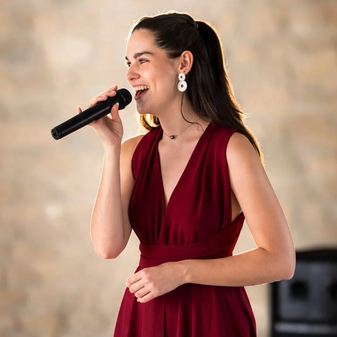 Une femme souriante, en robe rouge, chante dans un micro lors d'une performance ou d'un événement.