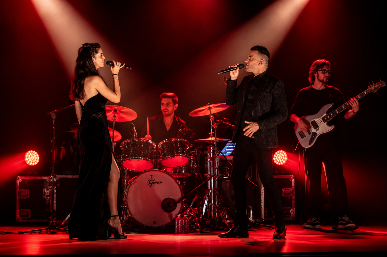 Un groupe de musiciens en performance sur scène, avec deux chanteurs, une femme en robe noire et un homme en costume noir, entourés de musiciens jouant de la batterie et de la basse, sous un éclairage rouge dramatique.
