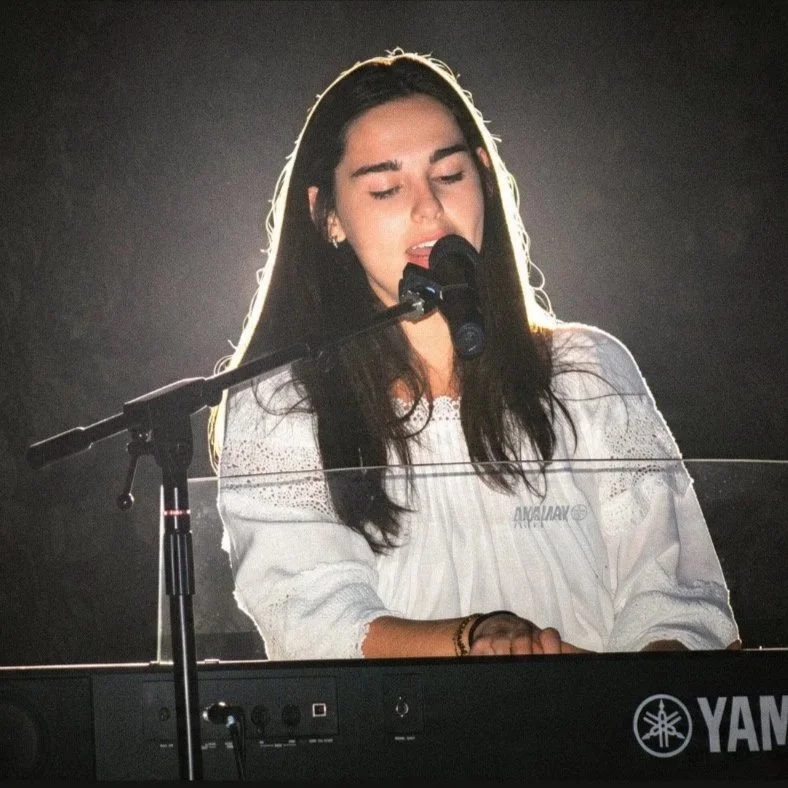 Une femme joue du piano ou du clavier Yamaha, avec un micro devant elle, les yeux fermés, probablement en train de chanter ou de jouer de la musique, dans un environnement sombre.