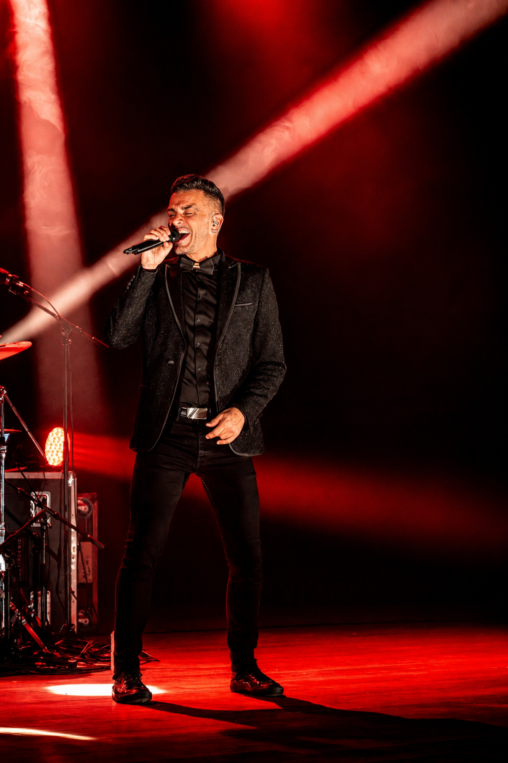 Un chanteur homme en costume noir chantant sur scène avec des lumières rouges et une audience invisible.