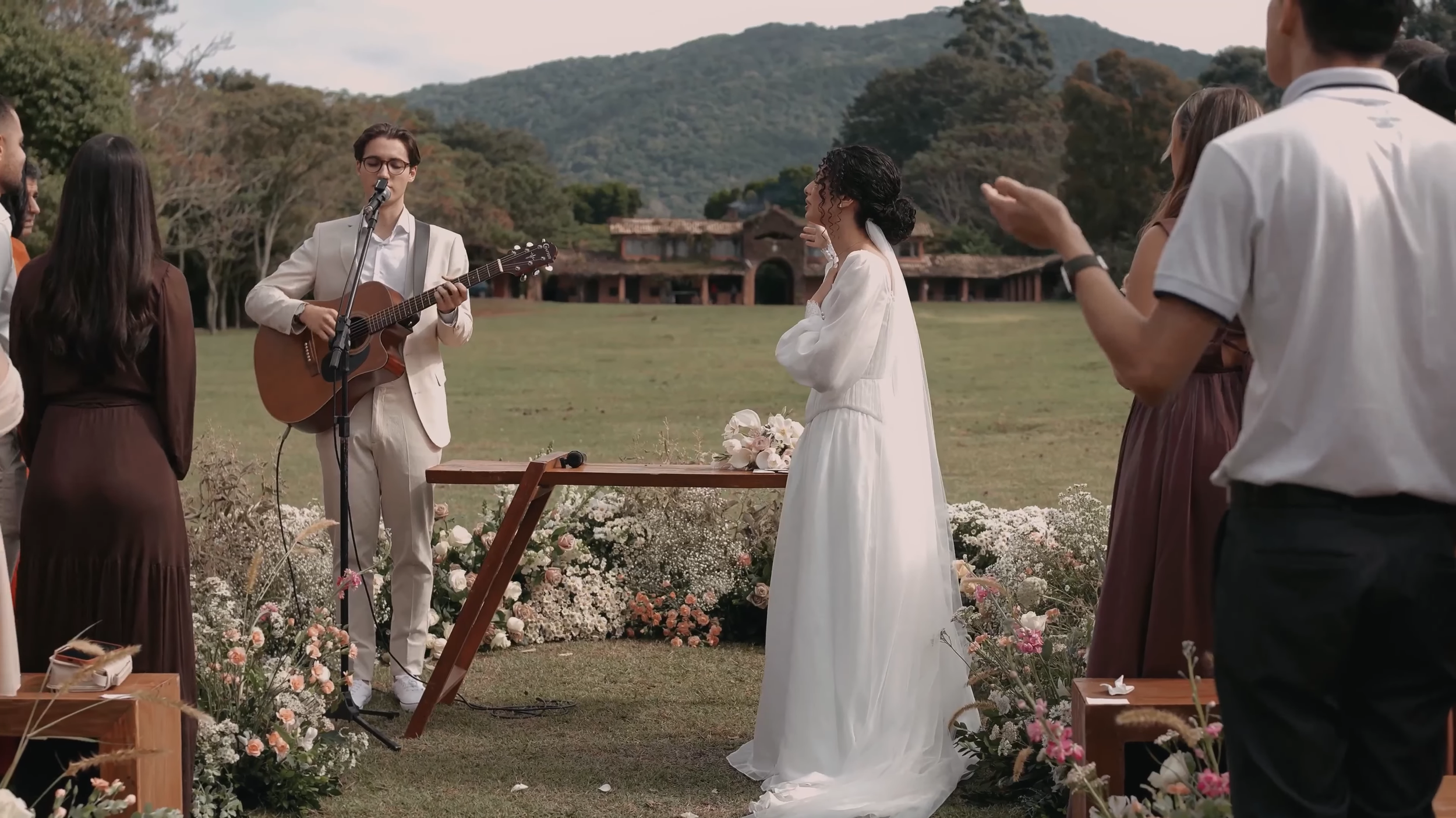 Un groupe de personnes assiste à un mariage en plein air. La mariée, habillée en robe blanche, se trouve devant un jeune homme qui joue de la guitare et chante, probablement lors de la cérémonie de mariage. Autour d'eux, il y a des invités et un décor floral avec des fleurs roses et blanches.