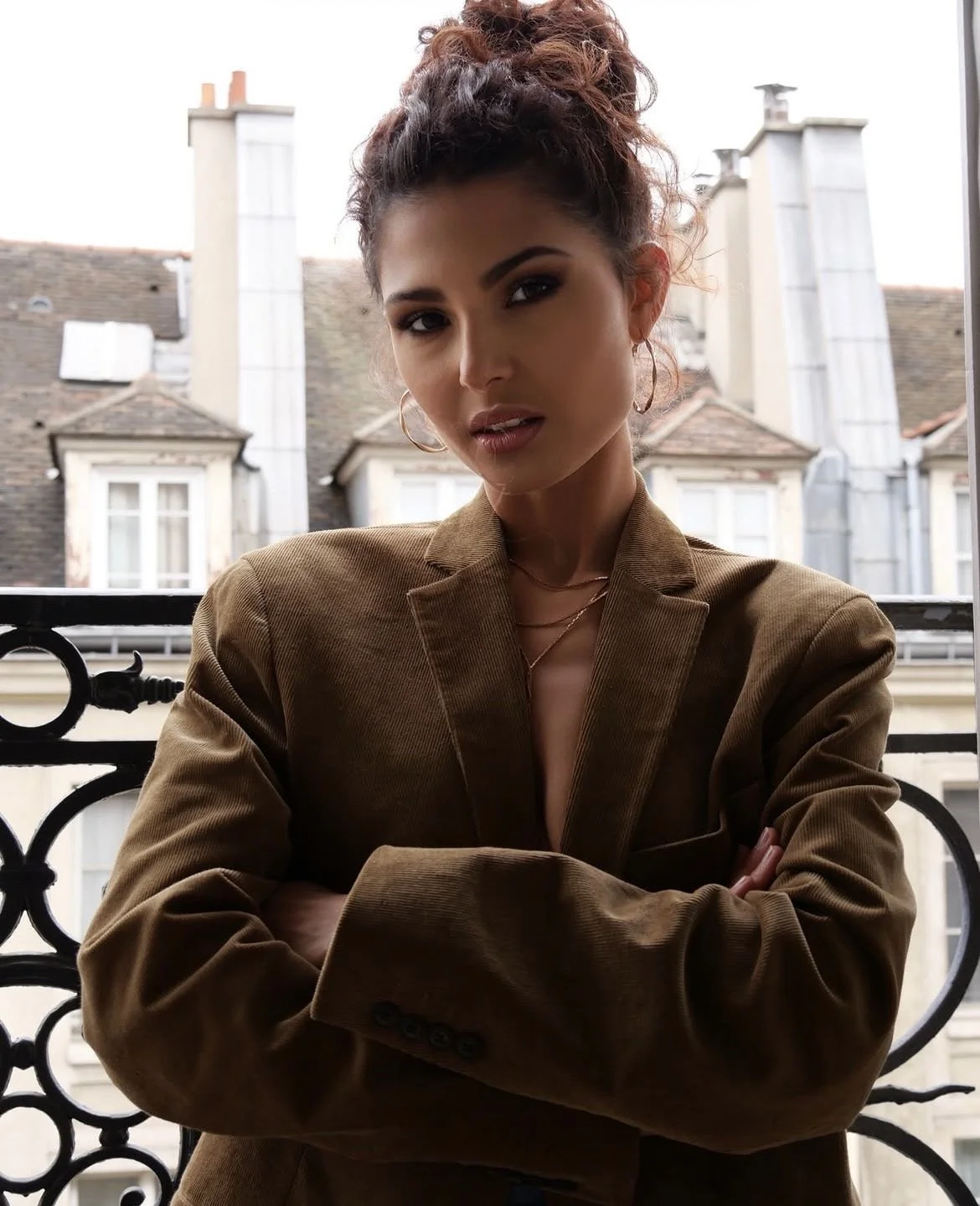 Une femme jeune et élégante pose sur un balcon avec un extérieur urbain de bâtiments en arrière-plan, portant un blazer marron et des bijoux en or, avec les bras croisés et un regard confiant.