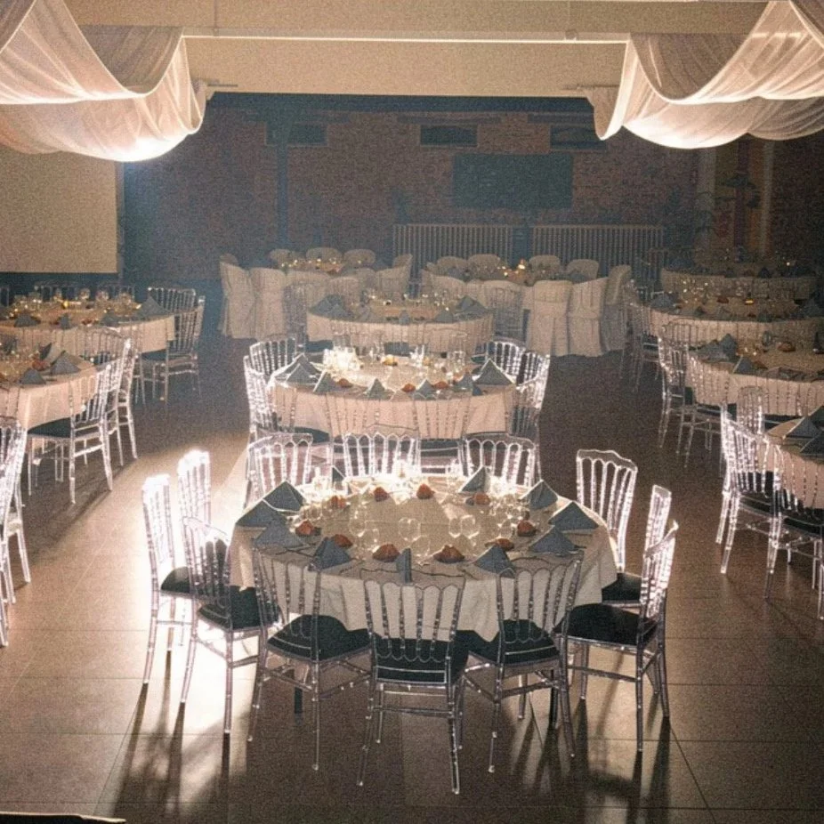 Salle de réception élégante avec plusieurs tables rondes décorées, lanternes, nappes blanches et chaises transparentes. Éclairage tamisé et drapés suspendus au plafond.
