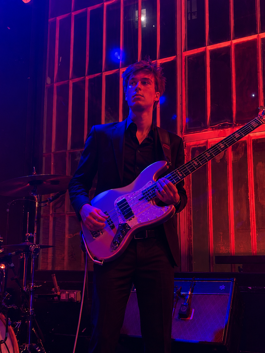 Un jeune homme joue de la basse électrique lors d'une performance dans un lieu sombre avec un éclairage rouge et violet, derrière une grande fenêtre en verre.