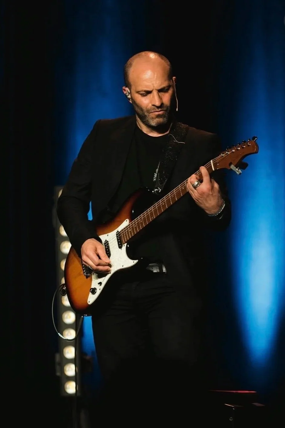 Un homme jouant de la guitare électrique sur scène avec un fond sombre et une lumière bleue.