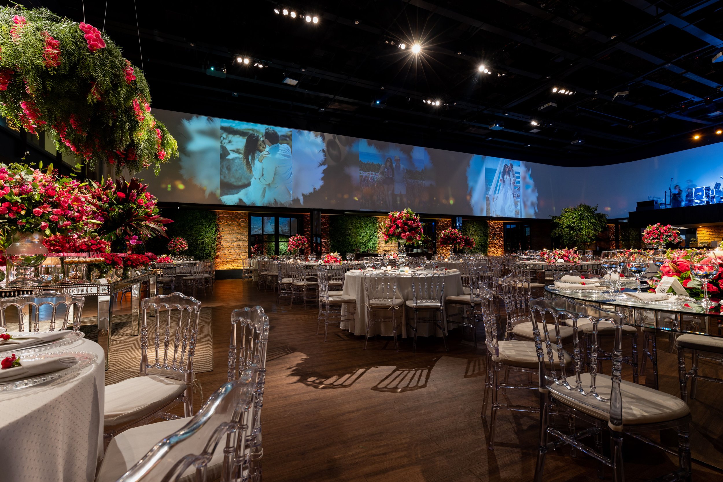 Salle de réception décorée avec de nombreuses fleurs roses et rouges, tables élégantes avec vaisselle et verres transparents, grandes écrans avec images de couples, ambiance festive et lumineuse.