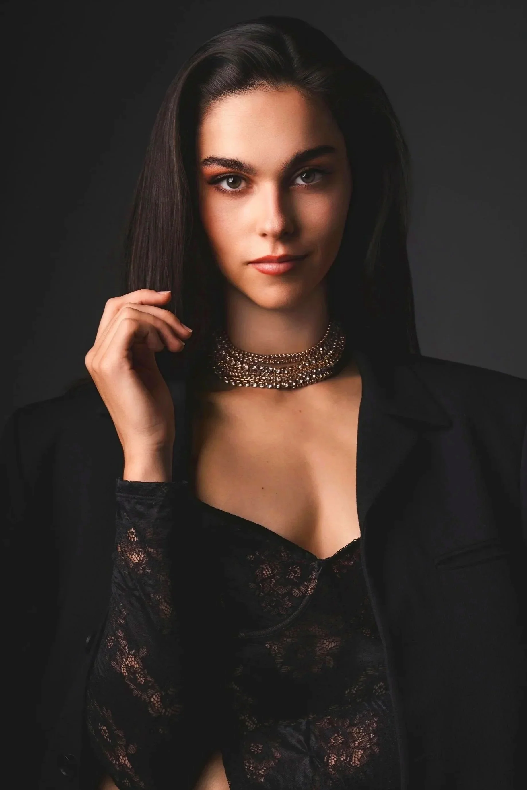 Jeune femme avec des cheveux longs, costume noir et collier or, posant contre un fond sombre.