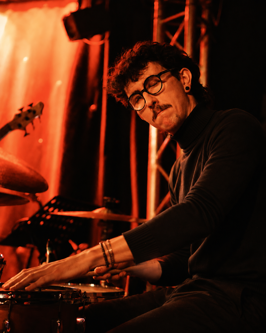 Jeune homme jouant du piano ou des percussions, portant des lunettes et un bracelet, dans un lieu avec un éclairage rouge, en train de se concentrer profondément.