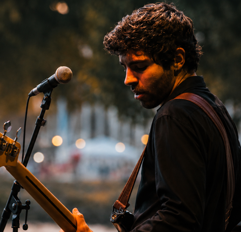 Un jeune homme joue de la guitare devant un microphone lors d'une performance en extérieur, avec un fond flou de arbres et de lumières