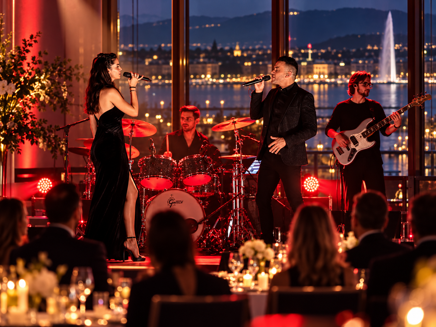 Un groupe de musiciens jouant en soirée dans un espace avec vue sur la ville de Genèvre et le jet d'eau, incluant une chanteuse, un chanteur, un batteur et un guitariste, lors d'un concert en intérieur avec décor élégant et éclairage chaud.